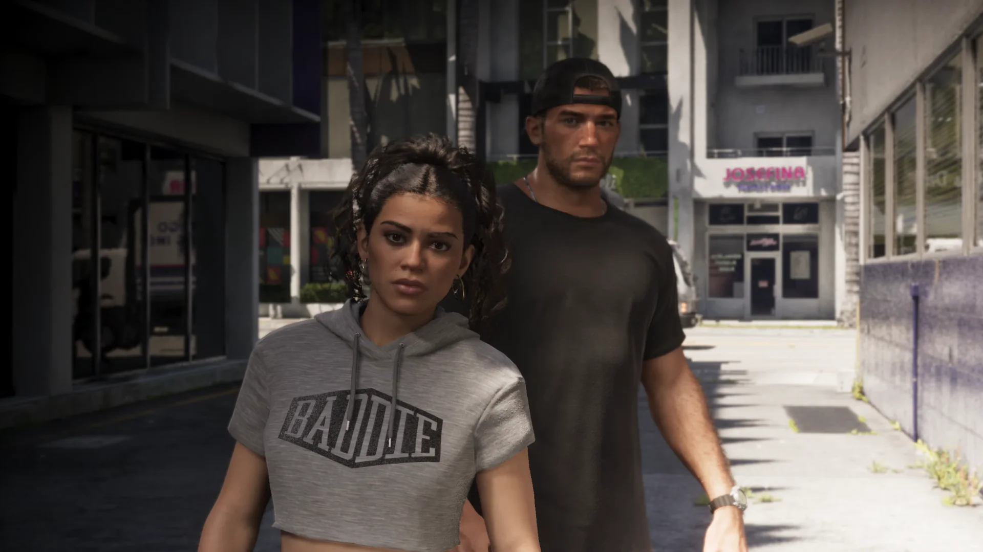 Lucia stands in a grey cropped hoodie while Jason stands behind her in a sunlit alley in Leonida.