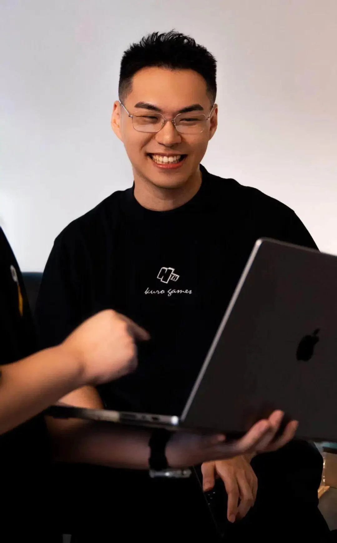 Solon Lee smiles while holding an open laptop and wearing a black Kuro Games shirt.